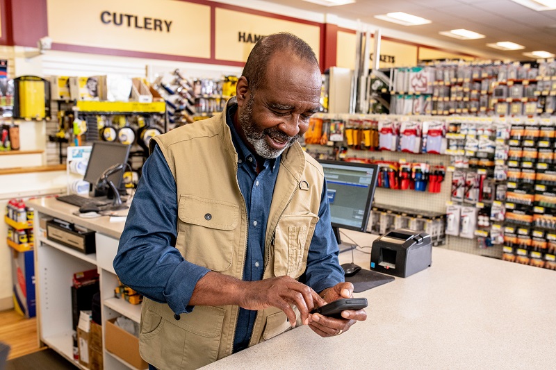 A man stands at the counter of his outdoor sporting goods store and views his business performance on his phone using the Monit app