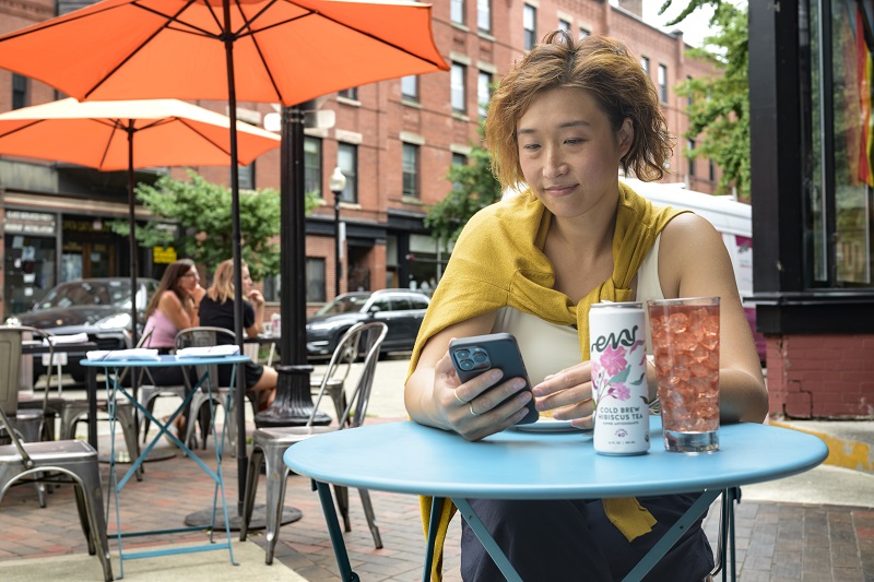 A woman sits outside at a small table and views her small business information on her phone in the Monit tool
