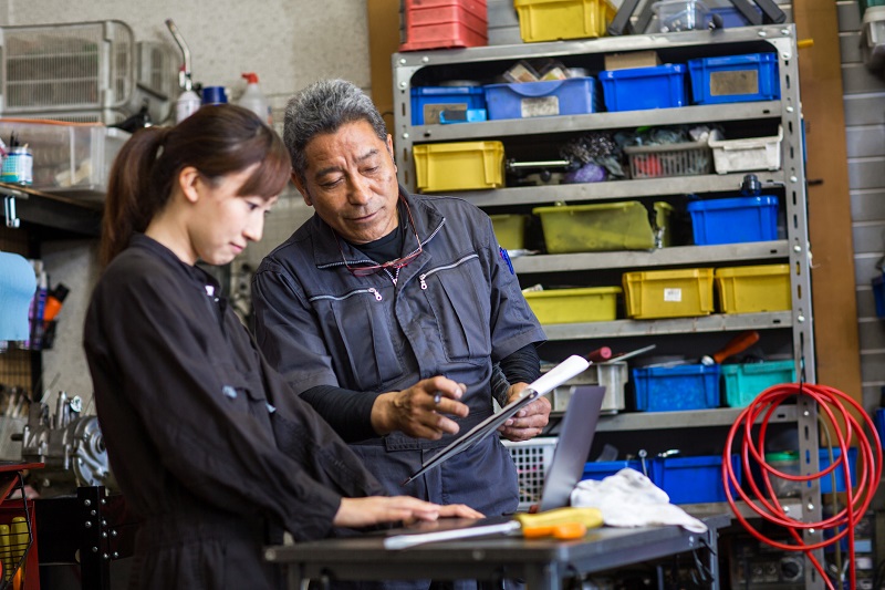 An autobody shop owner stands in his garage reviewing finances with an employee on a laptop