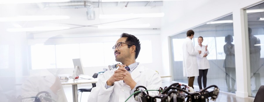 A scientist wearing a lab coat sits at his desk in his engineering lab reviewing his account on his computer