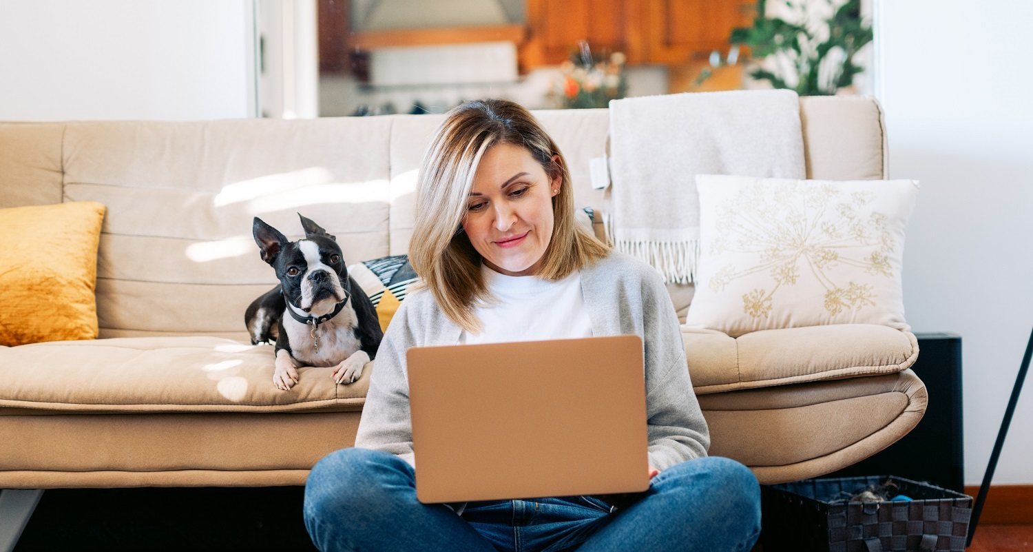 A woman sits with her dog while working on her laptop