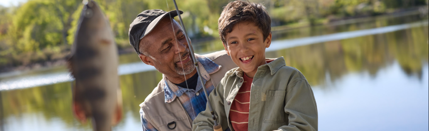 A boy proudly holds his fishing rod displaying his fish at the end while his grandfather sits beside him and smiles