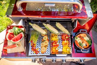 An open grill showcases a variety of grilled food including corn, peppers, salmon, and asparagus with a side of watermelon