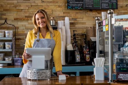 A woman stands at the checkout counter of her bakery while holding a computer tablet and smiling at the camera