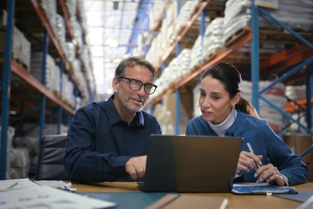 Two business partners sit together at a desk in a warehouse and review finances on a laptop