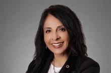 Beata wears a black blazer over a white top, has shoulder-length brown hair, and smiles at the camera